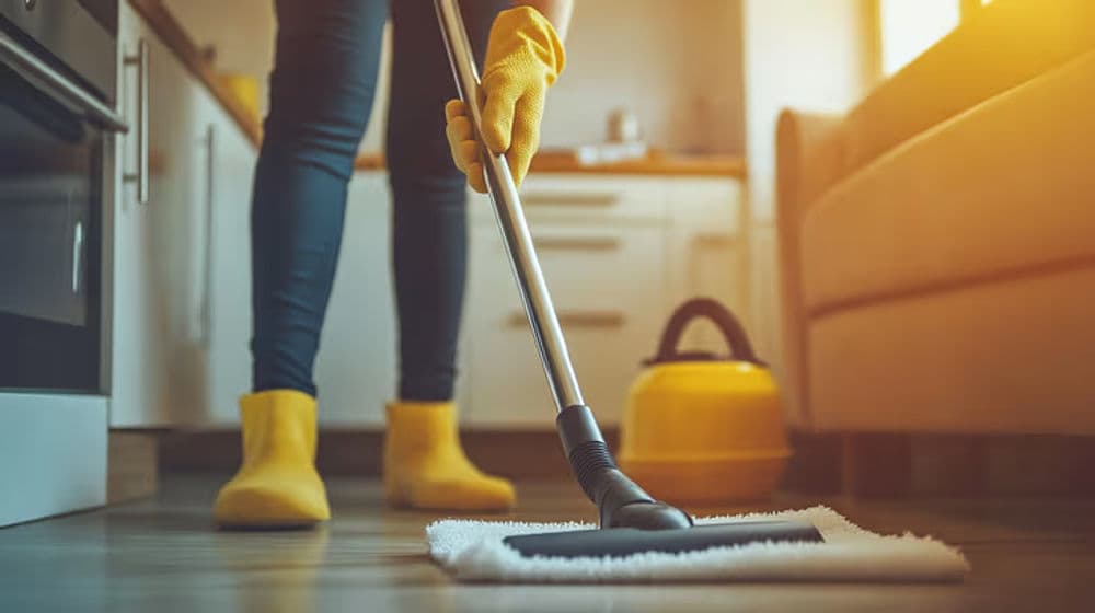 Person in yellow gloves mopping a kitchen floor with a vacuum cleaner nearby.